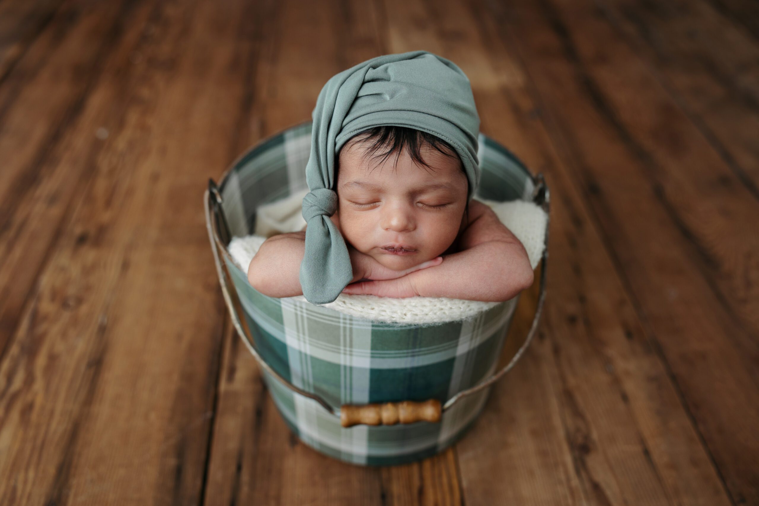 Newborn baby asleep in a cozy bucket, adorned with a cute bow hat, against a rustic wood backdrop.