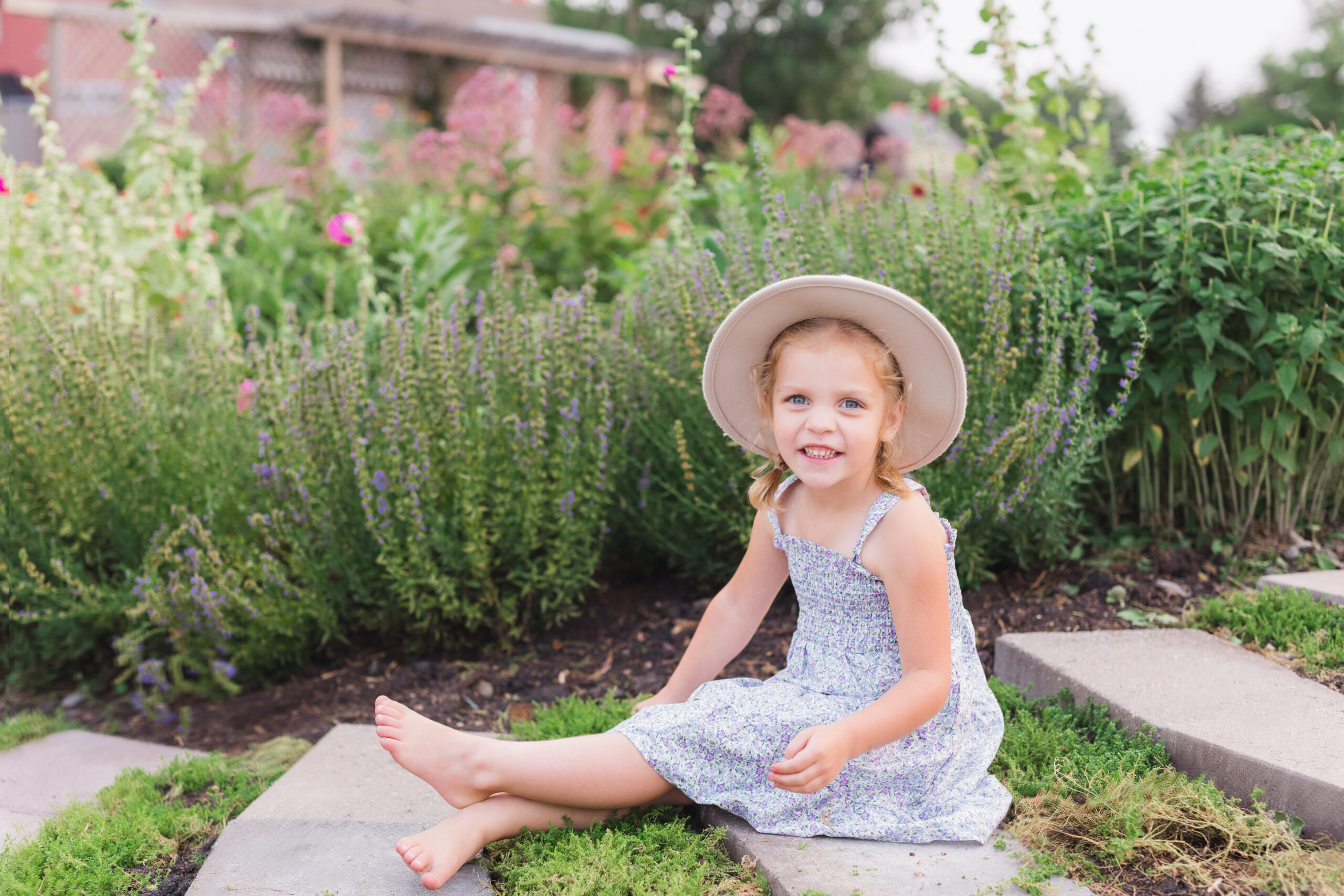 A young girl with a beaming smile, sitting in a lush garden, wearing a summer dress and a hat.