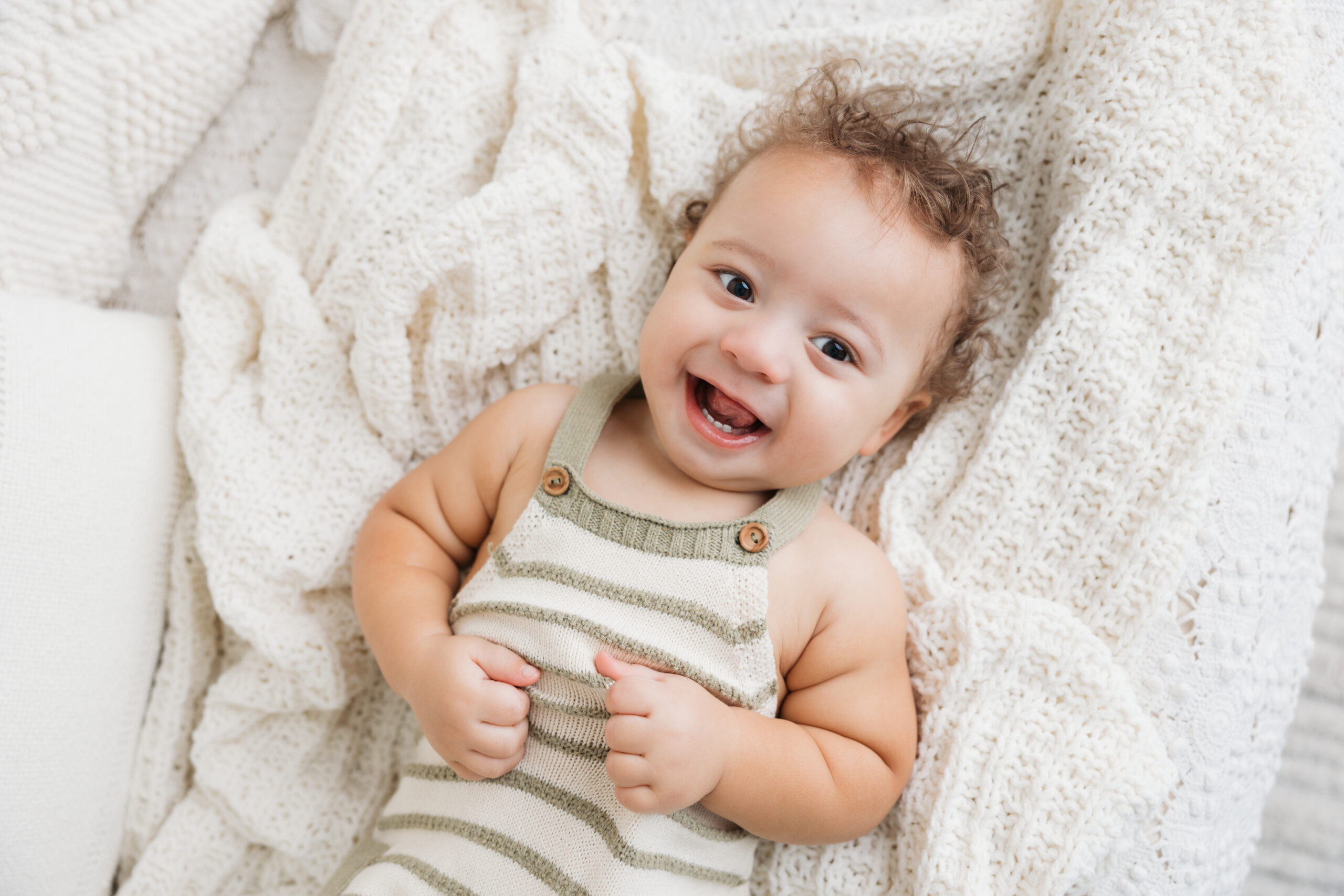A cheerful baby with curly hair, wearing a striped onesie, lies on a white knit blanket, laughing joyfully.