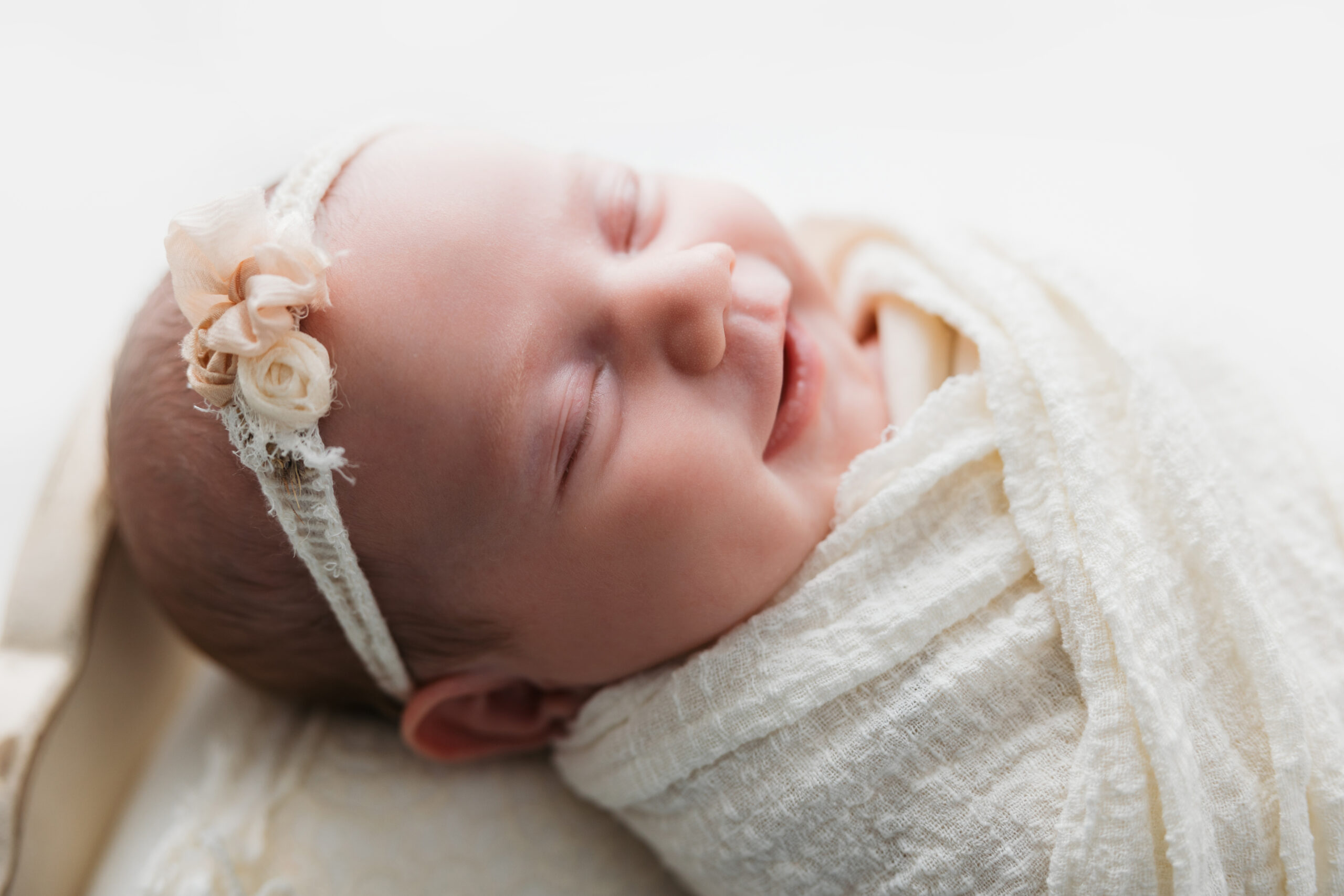 Newborn baby smiling in sleep, wrapped in a delicate cream swaddle with a floral headband
